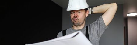 Young repairman in helmet, gloves and special uniform standing in empty flat, looking at construction plan and feeling frustrated over grey walls background. Professional repairman during work conceptの写真素材