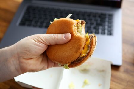 Close-up of male hand holding cheeseburger. Worker eating fast-food at workplace in office. Laptop...