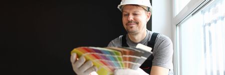 Young positive repairman in helmet and special uniform sitting in flat and choosing color for interior details over dark wall background. Professional repairman during work conceptの写真素材