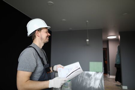 Young smiling repairman in helmet, gloves and special uniform standing in empty flat and holding...