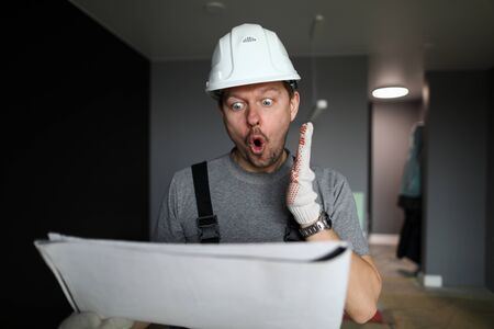 Young repairman in helmet, gloves and special uniform standing in empty flat, looking at...