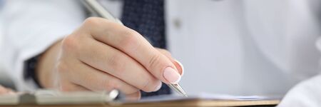 Close-up of professional female doctor writing prescription with silver pen. Red stethoscope on womans neck. Modern medicine and patient treatment conceptの写真素材