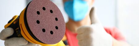 Close-up of craftsman holding electric sander. Protective mask on face and white gloves. Worker...