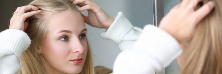 Portrait of young beautiful blonde woman looking in mirror. Pretty light-haired female with cute natural makeup holding hands near head. Modeling and beauty conceptの写真素材