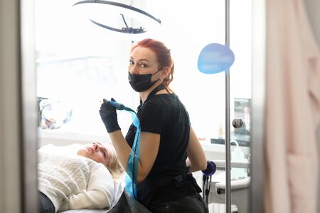 Portrait of cosmetologist working in salon. Young woman using special tool to apply permanent makeup...