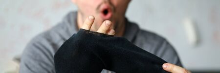 Man sitting at home surprised by big hole socks. Lack funds for purchase necessary things during economic crisis. Man put his fingers sock. Poverty against quarantine. Guy apartment in self-isolationの写真素材