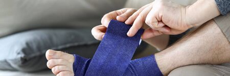 Man sits at home couch and bandages foot bandage. Uniform compression all areas for foot massage effect. Compensates for joint instability during movement. Increase blood circulation and relieve painの写真素材