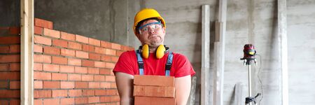 Portrait of foreman carrying stack of red bricks. Funny handyman making faces while working on...