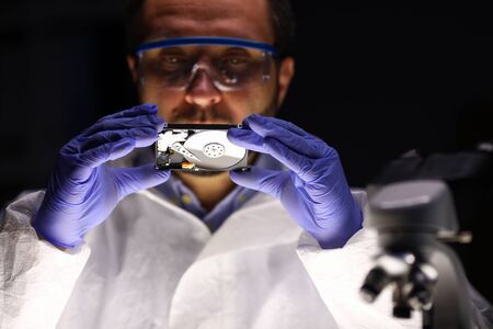 Portrait of specialist in eyewear looking at rigid disc. Man examining pc detail. Worker checking...