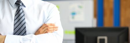 Manager in a tie stands in the office with his arms crossed. Man in business clothes on the background of office furnitureの写真素材