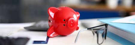 Close-up of red piggy bank laying on white desk. Container for saving money. Black stylish glasses. Modern design in office. Cash and investment for future conceptの写真素材