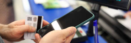 Close-up of male person paying for clothes via smartphone and plastic credit card. Cash register in...