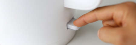Close-up of person pressing knob on electrical kettle with finger. Power switch on. Warm water for...
