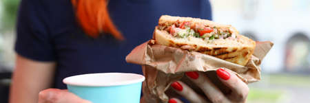Close-up of woman holding cup of hot coffee and delicious sandwich. Break for quick lunch. Female sitting outdoors on summer platform. Tasty brunch on fresh air. Food conceptの写真素材