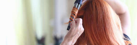 Close-up of red haired woman using curling iron for luxury hairstyle. Female preparing for festive event. Device to create beautiful hair at home. Technology and beauty conceptの写真素材