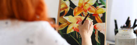 Close-up of talented female creating new picture of flowers. Creative red haired painter sitting in studio. Bright masterpiece of plants. Art and hobby conceptの写真素材