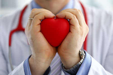 Male medicine doctor hands holding and covering red toy heart closeup. Cardio therapeutist student education physician make cardiac physical heart rate measure arrhythmia conceptの写真素材