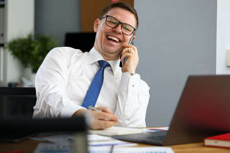 Portrait of businessman in good mood talking on phone with partner. Man in office sitting by desk with laptop and papers. Manager or secretary, boss or company directorの写真素材