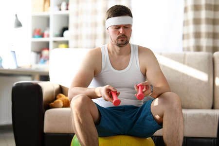 Young man sitting in an apartment on fitness ball. Man at home sits on ball in hands dumbbell. Training at home during quarantine. Physical training for office staff. Include fitness in your scheduleの写真素材