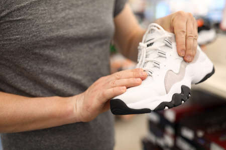 Close-up of persons hands holding stylish white sneakers. Male wearing grey shirt. Man choosing...