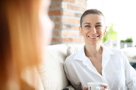 Portrait of charming female wearing white shirt and holding cup of coffee. Nice conversation with best friend. Spare time at home. Communication and relax on sofa conceptの写真素材