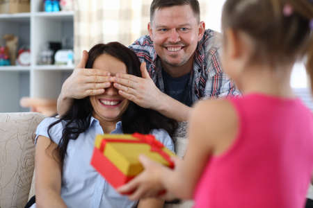 Portrait of smiling father closing mother eyes. Surprised woman waiting for present. Young pretty daughter giving gift to mom. Happy family and birthday conceptの写真素材