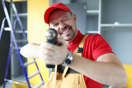 Portrait of cheerful handyman having fun on construction site. Man holding drill as gun. Good mood. Smiling funny worker. Cute uniform. Renovation and diy conceptの写真素材