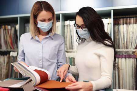 Two women wearing protective medical masks selecting fabric from catalog. New reality during covid19 pandemic conceptの写真素材