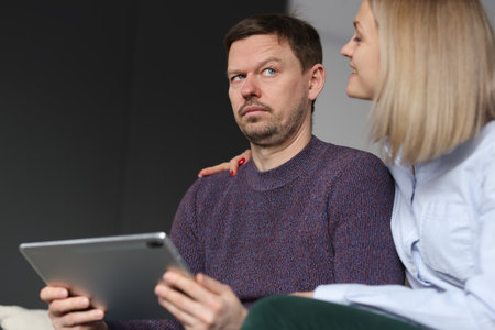 Pensive man sits with tablet next to smiling woman. Matriarchal marriage conceptの写真素材