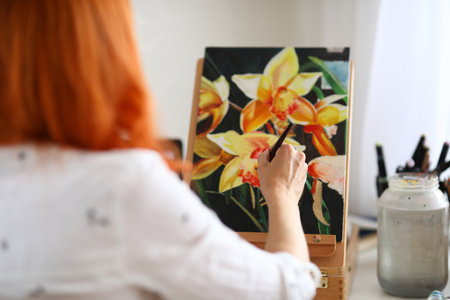 Close-up of talented female creating new picture of flowers. Creative red haired painter sitting in studio. Bright masterpiece of plants. Art and hobby conceptの写真素材