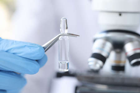 Hand in glove holds vial with vaccine against background of microscope. New vaccine production...