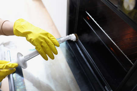 Female hands in yellow gloves are holding a steam cleaner and cleaning oven. Steam cleaner for home...