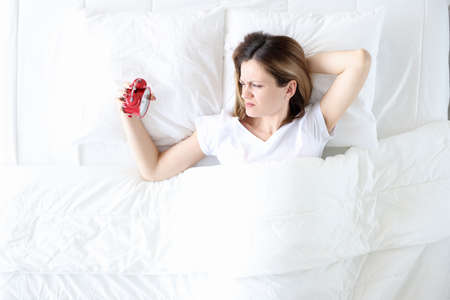 Young woman lying in bed and holding alarm clock in her hands top view