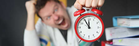 Man shouting and holding red alarm clock at table in office closeup