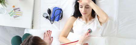 Doctor listening to woman patient with stethoscope in bed top view