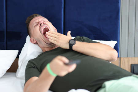 Young man lying in bed with remote control in his hand and yawning