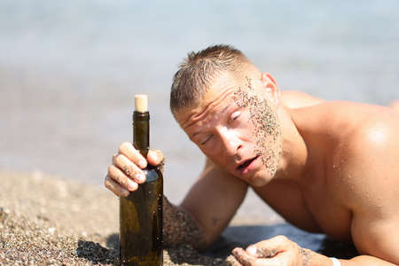 Drunk young man in sand lying on beach with bottle of wine in his handsの写真素材