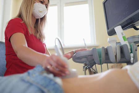 Doctor in medical protective mask doing ultrasound examination of abdominal cavity to patient in...