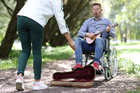 Disabled man in wheelchair plays guitar in park passers-by give money.の写真素材