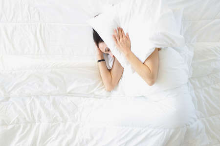 Young woman lying in white bed and covering her ears with pillow top viewの写真素材