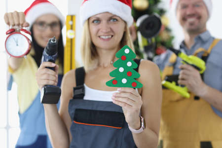 Group of foreman repairmen in santa claus hats holding drill and alarm clock near christmas tree closeupの写真素材