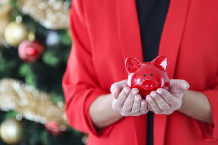 Woman in red jacket holds piggy bank against the background of Christmas tree.の写真素材