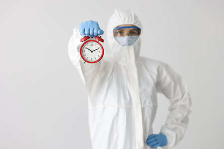 Woman in protective medical suit and mask with glasses holds red alarm clock on white backgroundの写真素材