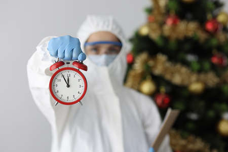 Woman in protective medical suit holds red alarm clock against background of Christmas treeの写真素材