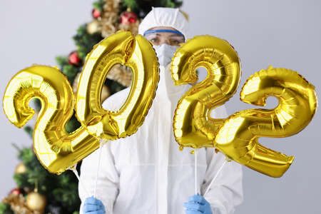 Woman in protective suit and mask holds numbers 2022 against background of Christmas tree