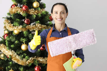 Portrait of young smiling woman holding thumbs up with mop and bucket on background of Christmas...