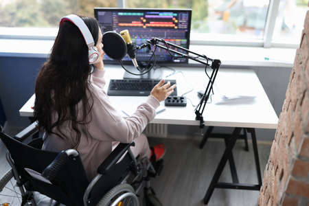 Woman in wheelchair with headphones speaks into microphone in front of computer monitorの写真素材