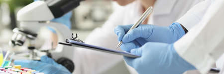 Woman chemist looking through microscope in laboratory. Assistant writing data to paper