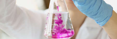 Chemist holds flask with clear liquid in hands and drops purple solvent into itの写真素材