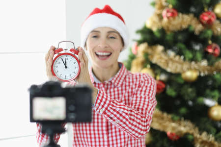 Joyful woman blogger shoots video on camera holding alarm clock on background of New Year treeの写真素材